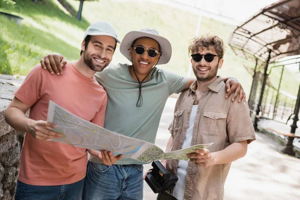 Беззаботный африканский американец в шляпе и солнцезащитных очках обнимает друзей на городской улице — стоковое фото