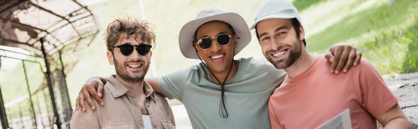 Carefree african american man in sunglasses embracing friends and smiling at camera on city street, banner — Stock Photo