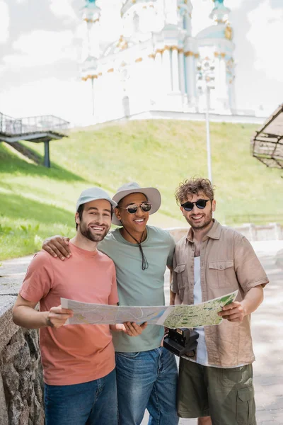 Щасливі багатонаціональні туристи у сонячних окулярах, які тримають карту міста і посміхаються біля церкви Сент-Ендрюс на розмитому фоні — стокове фото