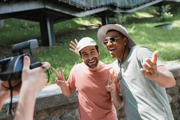 Blurred man with vintage camera taking photo of carefree interracial friends posing in sun hats and gesturing in city park — Stock Photo