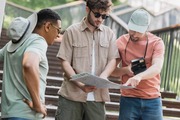 Man with vintage camera pointing at city map in hands of friend on stairs in city — Stock Photo