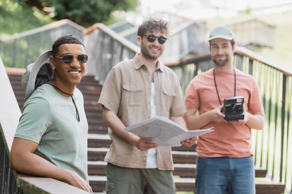 Молодой человек держит в руках карту и улыбается фотоаппарату рядом с друзьями-представителями разных рас на лестнице на улице города — стоковое фото