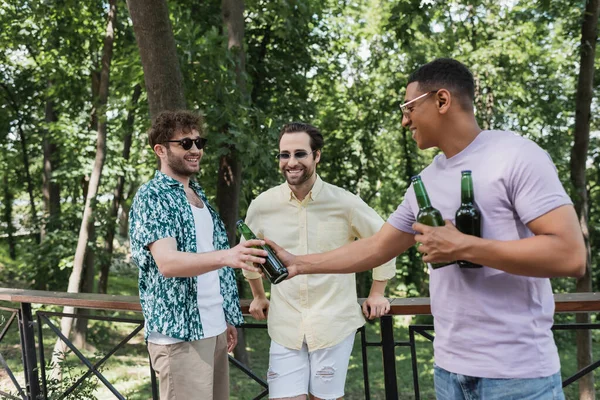 African american man giving fresh beer to happy friend near fence in city park — Stock Photo