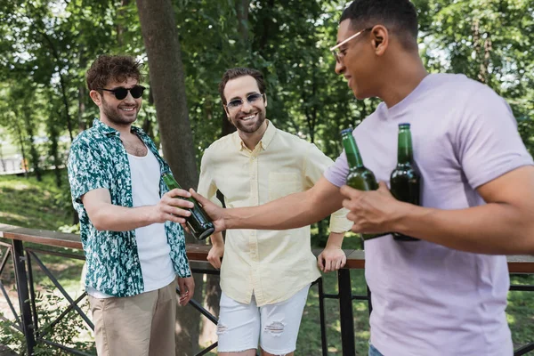 Homme afro-américain donner de la bière à des amis heureux dans des lunettes de soleil tout en passant du temps dans le parc de la ville — Photo de stock