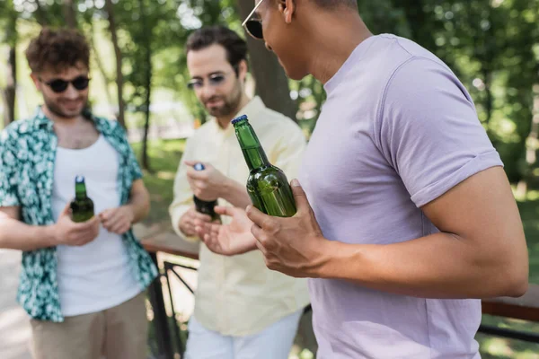 African american man holding bottle of fresh beer near trendy blurred friends in park — Stock Photo
