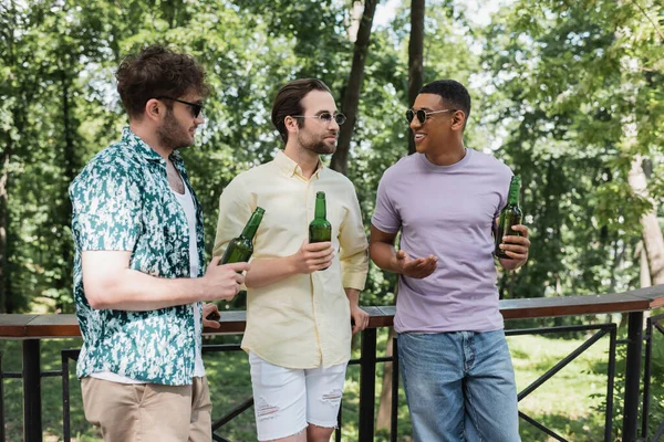 Happy and stylish multiethnic friends in sunglasses holding fresh beer and talking near fence in city park — Stock Photo