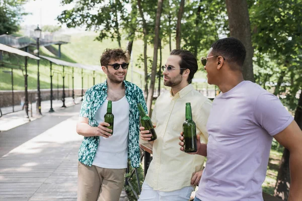Amigos inter-raciais despreocupados em roupa de verão elegante segurando garrafas de cerveja e falando no parque urbano — Fotografia de Stock