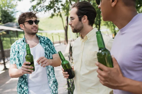 Hombre joven y de moda en gafas de sol sosteniendo cerveza y hablando con amigos interracial borrosa en el parque de la ciudad - foto de stock