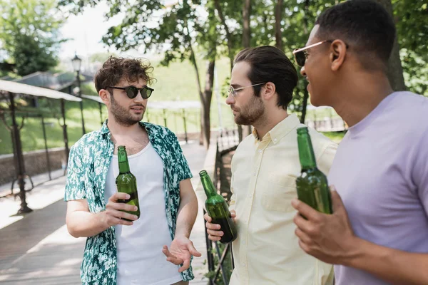Homem elegante em óculos de sol segurando cerveja e gesticulando enquanto conversa com amigos multiétnicos no parque da cidade no dia de verão — Fotografia de Stock