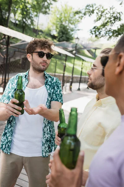 Молодий і модний чоловік в сонцезахисних окулярах вказує на пляшку пива біля розмитих міжрасових друзів в міському парку — стокове фото