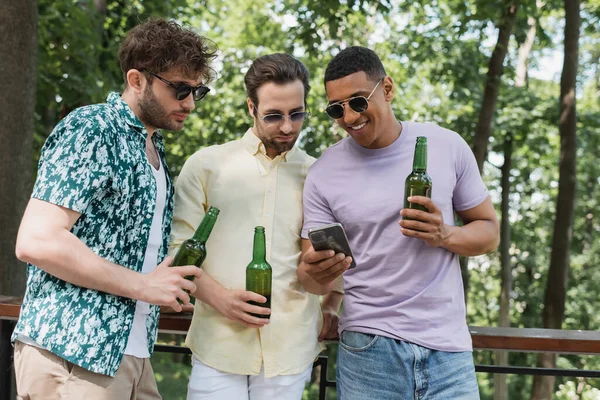 Alegre africano americano hombre en gafas de sol mostrando smartphone a amigos sosteniendo cerveza en parque - foto de stock