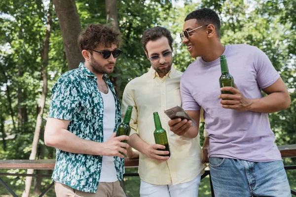 Homens elegantes segurando cerveja e olhando para o smartphone na mão de amigo americano africano feliz no parque da cidade — Fotografia de Stock