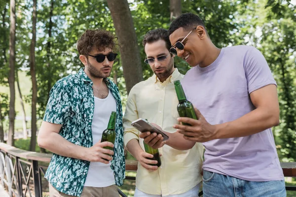 Alegre afroamericano hombre en gafas de sol mostrando el teléfono móvil a los amigos de moda sosteniendo cerveza en el parque de la ciudad - foto de stock
