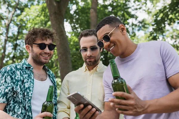 Улыбающийся африканский американец показывает мобильный телефон модным друзьям с пивными бутылками в зеленом парке — стоковое фото