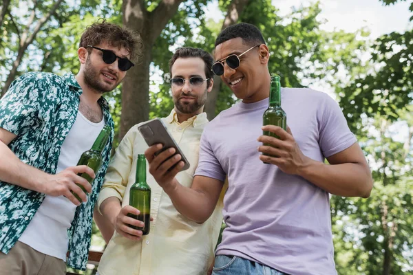Alegre afroamericano hombre sosteniendo el teléfono móvil cerca de elegantes amigos con botellas de cerveza en el parque - foto de stock