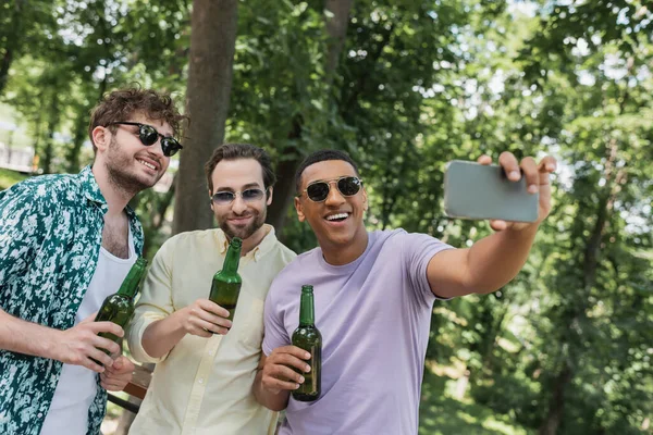 Hombre afroamericano sosteniendo cerveza y tomando selfie con amigos despreocupados en el parque de verano - foto de stock