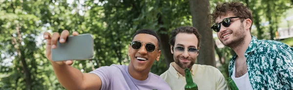 Carefree african american man in sunglasses taking selfie with trendy friends holding beer in park, banner — Stock Photo