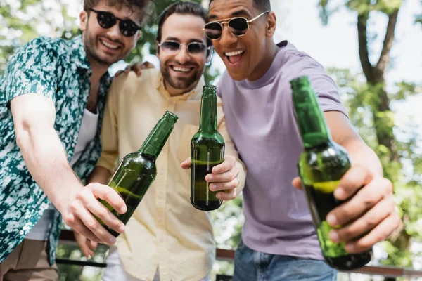 Blurred multiethnic men in stylish clothes and sunglasses toasting with beer in park — Stock Photo