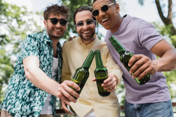 Беззаботные и стильные мультиэтнические мужчины в солнечных очках звенят пивными бутылками в парке — стоковое фото
