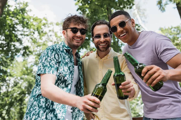 Amigos multiculturais alegres na moda roupas de verão e óculos de sol brindar com cerveja fresca no parque verde — Fotografia de Stock