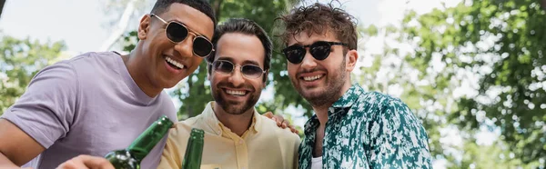Homens multiétnicos alegres e elegantes em óculos de sol sorrindo para a câmera perto de garrafas de cerveja no parque, banner — Fotografia de Stock