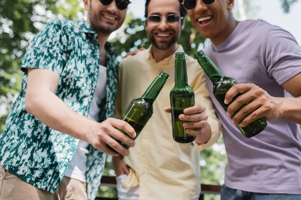 Selective focus of beer bottles in hands of carefree interracial friends spending time in summer park — Stock Photo