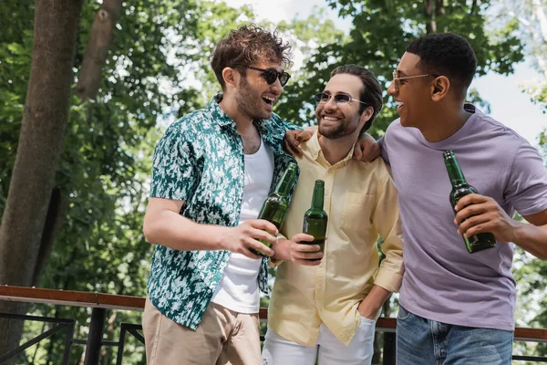 Amigos despreocupados multiculturales en traje de verano elegante abrazando mientras sostiene la cerveza en el parque de la ciudad - foto de stock