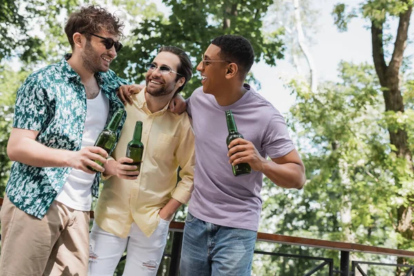 Amis interracial insouciants dans des lunettes de soleil et tenue d'été tendance embrassant tout en tenant des bouteilles de bière dans le parc — Photo de stock