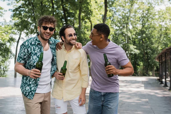 Carefree multiethnic friends in trendy sunglasses embracing while walking with beer in urban park — Stock Photo