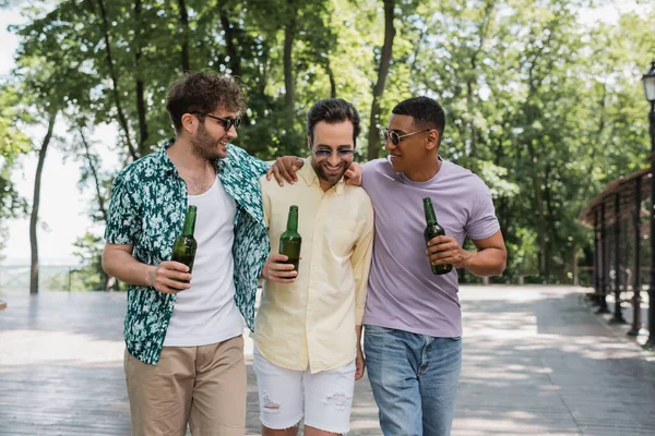 Amigos interracial despreocupados y elegantes abrazando y sosteniendo la cerveza durante el paseo de verano en el parque de la ciudad - foto de stock