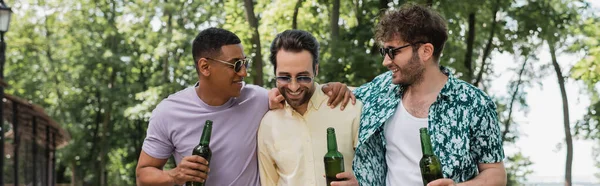 Amigos multiétnicos alegres e na moda segurando cerveja e abraçando enquanto passa o tempo no parque de verão, banner — Fotografia de Stock