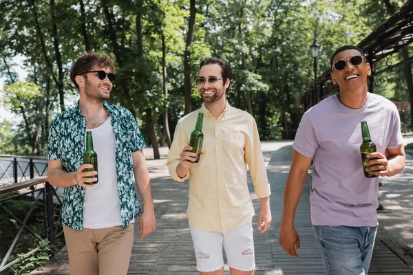 Весёлые и модные многонациональные друзья, держащие свежее пиво во время прогулки по городскому парку в летний день — стоковое фото
