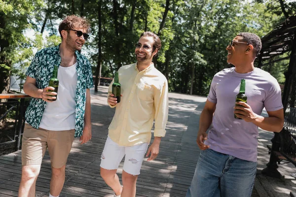Amigos despreocupados multiculturales en traje de verano de moda y gafas de sol caminando con cerveza en el parque urbano - foto de stock