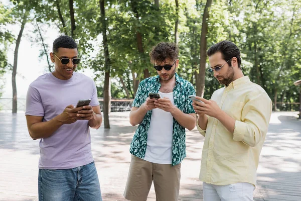 Homme afro-américain joyeux dans des lunettes de soleil en utilisant le téléphone mobile avec des amis élégants dans le parc de la ville — Photo de stock