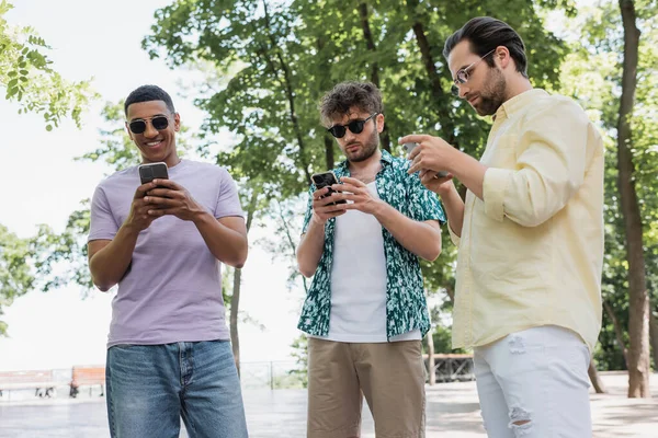 Multicultural men in stylish summer outfit and sunglasses using smartphones in city park — Stock Photo