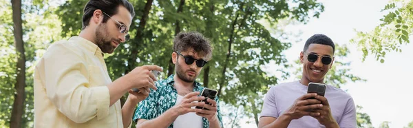 Беззаботный африканский американец с помощью смартфона рядом с друзьями в модных солнцезащитных очках в зеленом парке, баннер — стоковое фото
