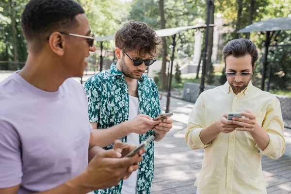 Afro-américain homme regardant des amis à la mode bavarder sur les téléphones mobiles dans le parc d'été — Photo de stock