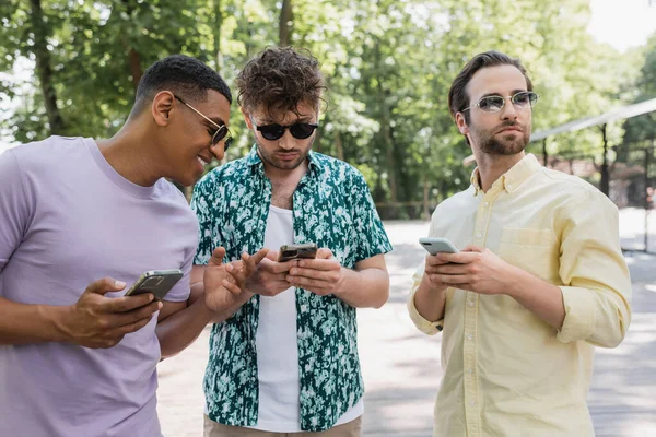 Joyeux homme afro-américain pointant du doigt près des amis à la mode en utilisant des téléphones cellulaires dans le parc d'été — Photo de stock