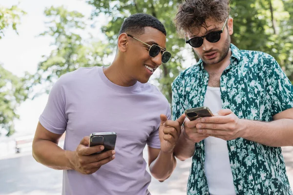 Sonriente afroamericano hombre apuntando con el dedo al teléfono inteligente en manos de un amigo de moda en el parque de verano - foto de stock