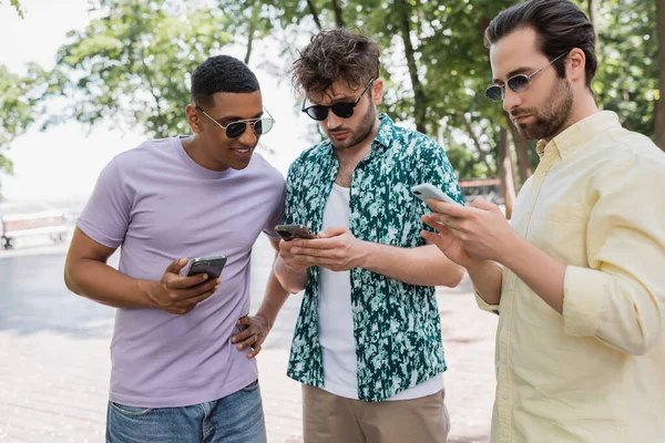 Sonriente hombre afroamericano en gafas de sol mirando a amigos usando teléfonos móviles en el parque urbano - foto de stock