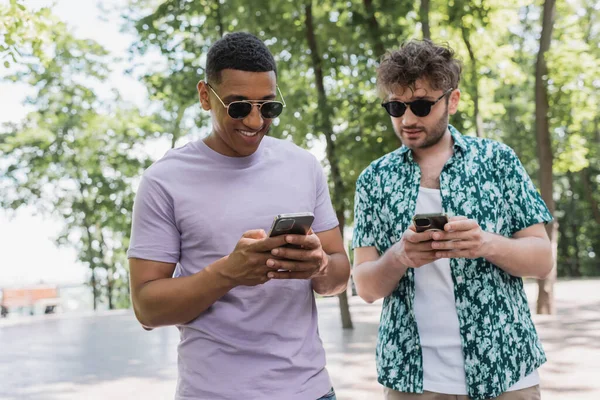 Улыбающиеся межрасовые друзья в солнечных очках с помощью смартфонов в парке — стоковое фото