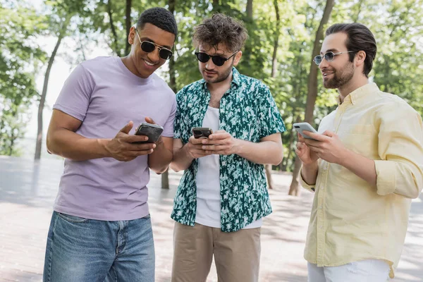 Un homme afro-américain positif utilisant un smartphone avec des amis proches dans des lunettes de soleil dans le parc — Photo de stock