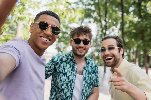 Amigos inter-raciais positivos em óculos de sol apontando para câmera no parque de verão — Fotografia de Stock