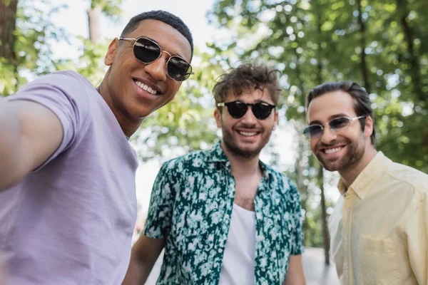 Homme afro-américain joyeux dans des lunettes de soleil debout près des amis flous dans le parc d'été — Photo de stock