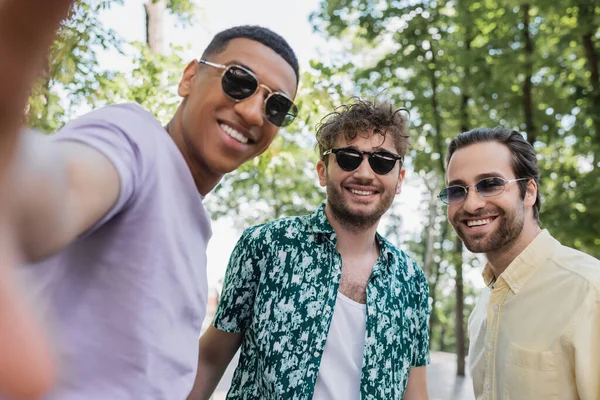 Alegre amigos multiétnicos en gafas de sol de pie en el parque en verano - foto de stock