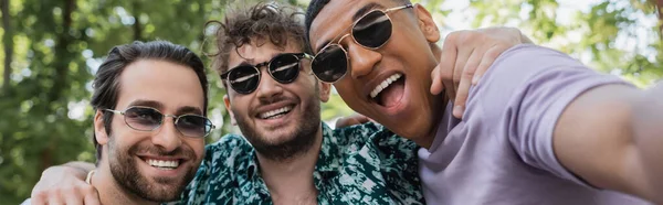 Alegres amigos multiétnicos en gafas de sol abrazándose en el parque de verano, pancarta - foto de stock