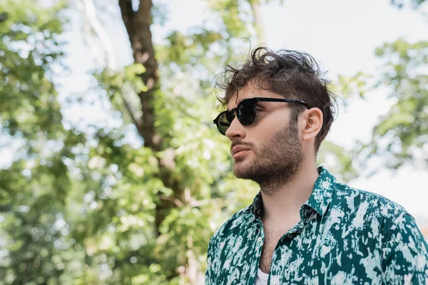 Молодий чоловік в сонцезахисних окулярах і сорочці стоїть в розмитому літньому парку — стокове фото