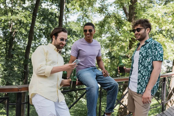 Cheerful interracial men with beer using smartphone in summer park — Stock Photo