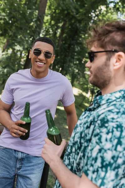Улыбающийся африканский американец в солнечных очках держит пиво рядом с размытым другом в летнем парке — стоковое фото