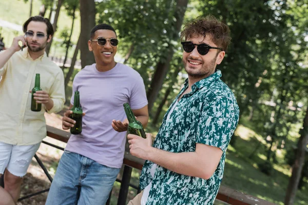 Веселый человек в солнечных очках, стоящий рядом с межрасовыми друзьями с пивом в летнем парке — стоковое фото